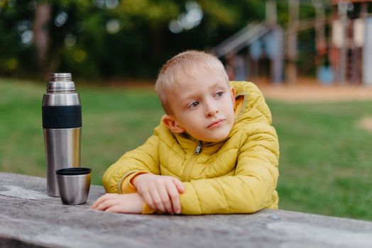 Boy is sitting at wooden table with cup of tea and looking into distance. thermos tea A boy sits at a table in the park in the fall season. Child Boy Son In Autumn Park, Sitting On Wooden Bench And Table. Little Kid Outdoors.