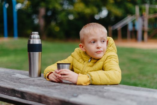 Boy is sitting at wooden table with cup of tea and looking into distance. thermos tea A boy sits at a table in the park in the fall season. Child Boy Son In Autumn Park, Sitting On Wooden Bench And Table. Little Kid Outdoors.