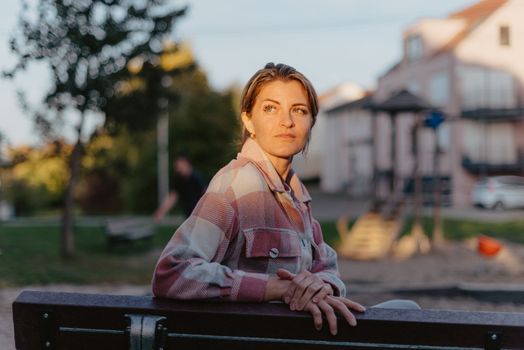 Girl sitting on a bench and watching distant city scenic. Girl enjoying city view from a bench in sunset or sunrise time. Beautiful blond-haired pretty girl in summer green park against sunset beams. Lonely woman sitting alone moments sunset. On Blurred background bokeh. light Fair.