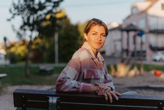 Girl sitting on a bench and watching distant city scenic. Girl enjoying city view from a bench in sunset or sunrise time. Beautiful blond-haired pretty girl in summer green park against sunset beams. Lonely woman sitting alone moments sunset. On Blurred background bokeh. light Fair.