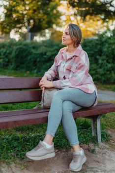 Girl sitting on a bench and watching distant city scenic. Girl enjoying city view from a bench in sunset or sunrise time. Beautiful blond-haired pretty girl in summer green park against sunset beams. Lonely woman sitting alone moments sunset. On Blurred background bokeh. light Fair.