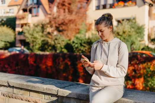 Young fashionable teenage girl with smartphone in park in autumn sitting at smiling. Trendy young woman in fall in park texting. Retouched, vibrant colors. Beautiful blonde teenage girl wearing casual modern autumn outfit sitting in park in autumn. Retouched, vibrant colors, brownish tones.