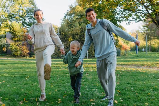 a family mom and 2 sons are having fun, running in the park and jumping. Sons, children, mom, run, play, rejoice, enjoy nature in summer. Family teamwork. Happy family team, running together in field, happily waving their hands. Group of people of different ages. happy family, mom and two sons on an emotional walk. Running and enjoying life in a green field in the park, blue sky, nature.