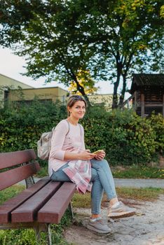 Girl sitting on a bench and watching distant city scenic. Girl enjoying city view from a bench in sunset or sunrise time. Beautiful blond-haired pretty girl in summer green park against sunset beams. Lonely woman sitting alone moments sunset. On Blurred background bokeh. light Fair.