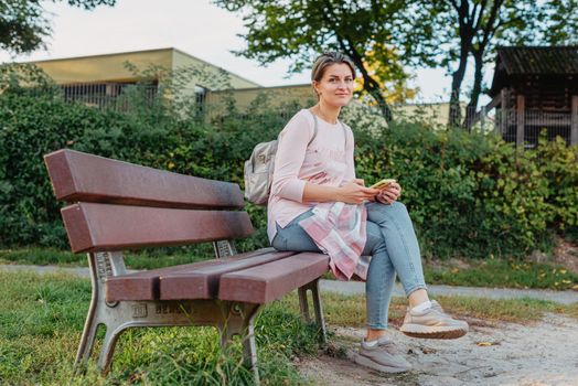 Girl sitting on a bench and watching distant city scenic. Girl enjoying city view from a bench in sunset or sunrise time. Beautiful blond-haired pretty girl in summer green park against sunset beams. Lonely woman sitting alone moments sunset. On Blurred background bokeh. light Fair.