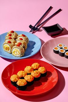 Delicious warm sushi rolls with tempura shrimp, maki sushi with creamy masago roe topping and salmon filling served on colorful plates with soy sauce on pink background. Popular Japanese cuisine