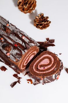 Sliced chocolate Swiss cake roll in shape of log filled with delicate berry custard cream garnished with dark chocolate flakes and candied strawberry pieces on white surface with gilded fir cones