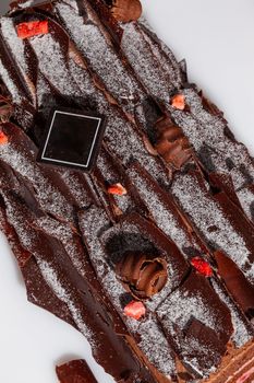 Log shaped cake roll covered with bark from thin dark chocolate shavings sprinkled with powdered sugar and pieces of candied strawberries on white background. Traditional Yule log. Festive treats