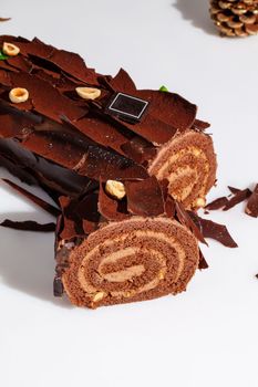 Sliced soft Swiss roll with buttercream filling covered with dark chocolate shavings sprinkled with cocoa and hazelnuts on white background with fir cones. Traditional New Year treats