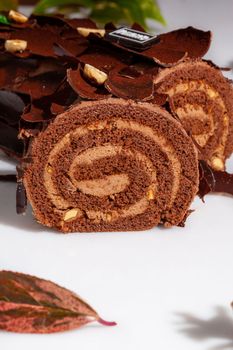 Closeup of sliced log-shaped sweet chocolate roll with tender buttercream filling, decorated with chocolate chips and hazelnuts on white background. Popular pastries concept