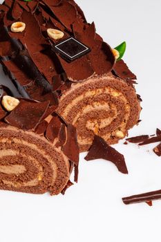 Sliced soft Swiss roll covered with dark chocolate shavings sprinkled with cocoa and hazelnuts, with delicate filling of buttercream with dried apricots and nuts on white background. New Year treats