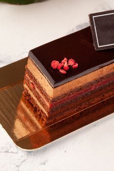 Delicate moist chocolate sponge cake with airy chocolate mousse and raspberry confit interlayer topped with glaze and candied berries served on golden cardboard on white background