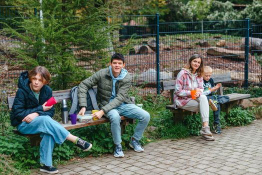 Happy Family drinking tea from a thermos in the forest. Photo of cute charming mother with childrens dressed casual outfit walking sitting bench drinking tea smiling outside city park