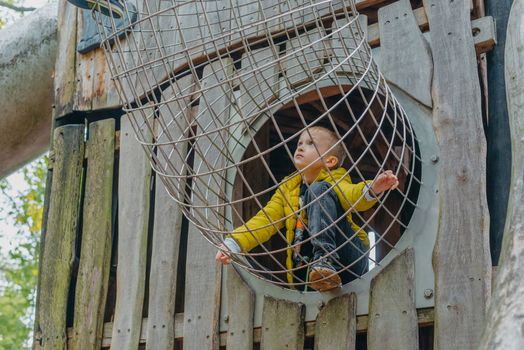 A child climbs up an alpine grid in a park on a playground on a hot summer day. children's playground in a public park, entertainment and recreation for children, mountaineering training. Child playing on outdoor playground. Kids play on school or kindergarten yard. Active kid on colorful slide and swing. Healthy summer activity for children. Little boy climbing outdoors.