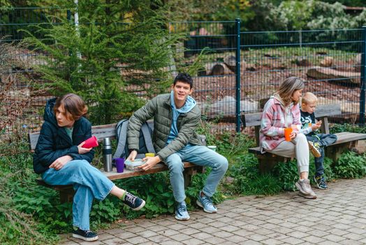 Happy Family drinking tea from a thermos in the forest. Photo of cute charming mother with childrens dressed casual outfit walking sitting bench drinking tea smiling outside city park