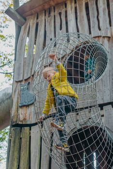 A child climbs up an alpine grid in a park on a playground on a hot summer day. children's playground in a public park, entertainment and recreation for children, mountaineering training. Child playing on outdoor playground. Kids play on school or kindergarten yard. Active kid on colorful slide and swing. Healthy summer activity for children. Little boy climbing outdoors.