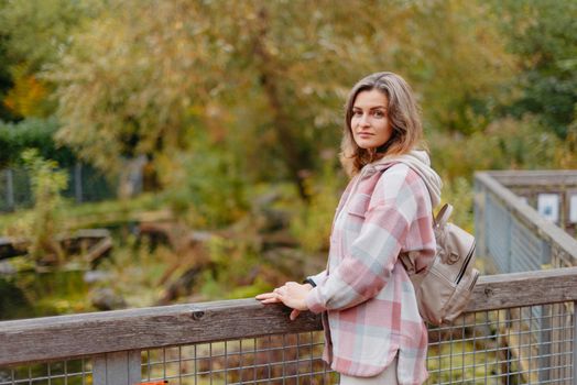 Portrait of cute young woman in casual wear in autumn, standing on bridge against background of an autumn Park and river. Pretty female walking in Park in golden fall. Copy space. smiling girl in the park standing on wooden bridge and looking at the camera in autumn season