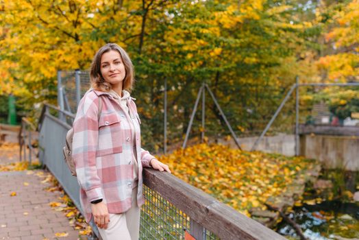 Portrait of cute young woman in casual wear in autumn, standing on bridge against background of an autumn Park and river. Pretty female walking in Park in golden fall. Copy space. smiling girl in the park standing on wooden bridge and looking at the camera in autumn season