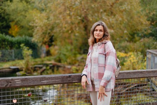 Portrait of cute young woman in casual wear in autumn, standing on bridge against background of an autumn Park and river. Pretty female walking in Park in golden fall. Copy space. smiling girl in the park standing on wooden bridge and looking at the camera in autumn season