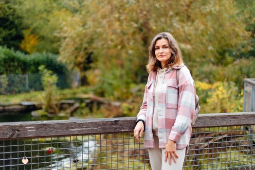 Portrait of cute young woman in casual wear in autumn, standing on bridge against background of an autumn Park and river. Pretty female walking in Park in golden fall. Copy space. smiling girl in the park standing on wooden bridge and looking at the camera in autumn season