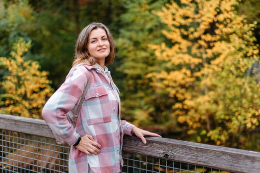 Portrait of cute young woman in casual wear in autumn, standing on bridge against background of an autumn Park and river. Pretty female walking in Park in golden fall. Copy space. smiling girl in the park standing on wooden bridge and looking at the camera in autumn season