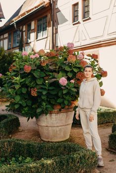 Attractive curly blonde woman walk on the city park street. Girl wear purple hoodie look happy and smiles. Woman make here me gesture standing near pink blooming bush flowers. Happy laughing girl