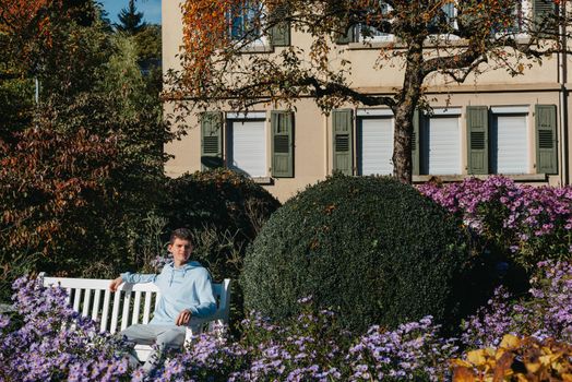 a teenager sits on a bench in the park drinks coffee from a thermo mug and looks into a phone. Portrait of handsome cheerful guy sitting on bench fresh air using device browsing media smm drinking latte urban outside outdoor.