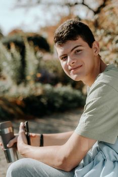 a teenager sits on a bench in the park drinks coffee from a thermo mug and looks into a phone. Portrait of handsome cheerful guy sitting on bench fresh air using device browsing media smm drinking latte urban outside outdoor.