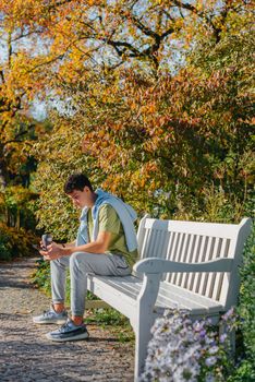 a teenager sits on a bench in the park drinks coffee from a thermo mug and looks into a phone. Portrait of handsome cheerful guy sitting on bench fresh air using device browsing media smm drinking latte urban outside outdoor.