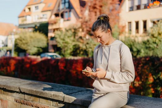 Young fashionable teenage girl with smartphone in park in autumn sitting at smiling. Trendy young woman in fall in park texting. Retouched, vibrant colors. Beautiful blonde teenage girl wearing casual modern autumn outfit sitting in park in autumn. Retouched, vibrant colors, brownish tones.