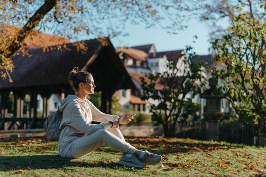 Young fashionable teenage girl with smartphone in park in autumn sitting at smiling. Trendy young woman in fall in park texting. Retouched, vibrant colors. Beautiful blonde teenage girl wearing casual modern autumn outfit sitting in park in autumn. Retouched, vibrant colors, brownish tones.