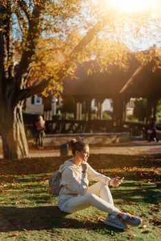 Young fashionable teenage girl with smartphone in park in autumn sitting at smiling. Trendy young woman in fall in park texting. Retouched, vibrant colors. Beautiful blonde teenage girl wearing casual modern autumn outfit sitting in park in autumn. Retouched, vibrant colors, brownish tones.