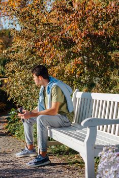 a teenager sits on a bench in the park drinks coffee from a thermo mug and looks into a phone. Portrait of handsome cheerful guy sitting on bench fresh air using device browsing media smm drinking latte urban outside outdoor.