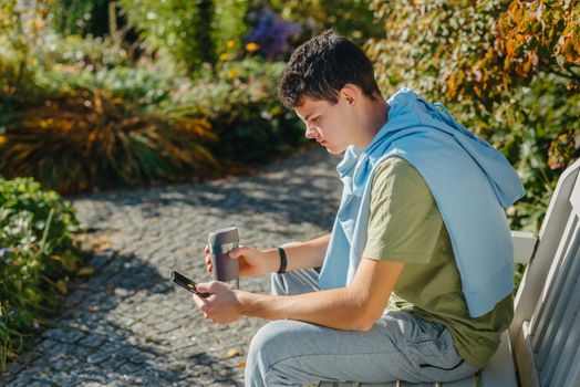 a teenager sits on a bench in the park drinks coffee from a thermo mug and looks into a phone. Portrait of handsome cheerful guy sitting on bench fresh air using device browsing media smm drinking latte urban outside outdoor.