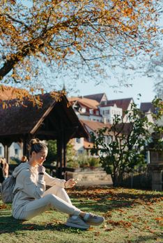 Young fashionable teenage girl with smartphone in park in autumn sitting at smiling. Trendy young woman in fall in park texting. Retouched, vibrant colors. Beautiful blonde teenage girl wearing casual modern autumn outfit sitting in park in autumn. Retouched, vibrant colors, brownish tones.