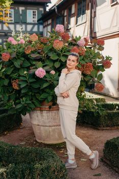 Attractive curly blonde woman walk on the city park street. Girl wear purple hoodie look happy and smiles. Woman make here me gesture standing near pink blooming bush flowers. Happy laughing girl