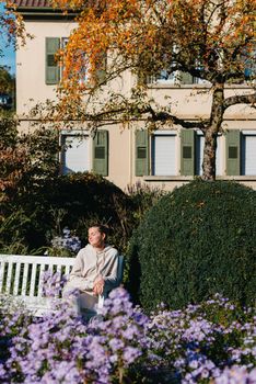 a girl sits on a bench in the park and enjoys the sun. Portrait young adult attractive woman enjoy sitting on bench and relaxing calm carefree rest in city park against green grass and trees on sunny day. Single female person relaxing chilling outdoors