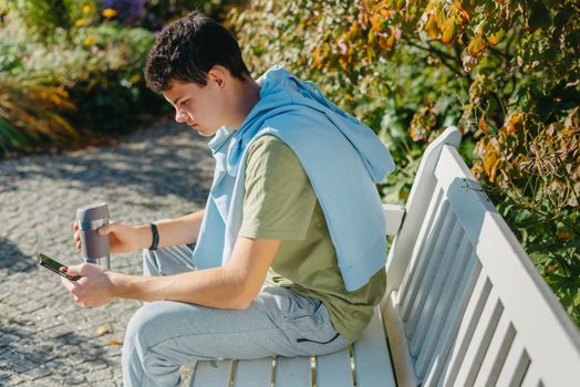 a teenager sits on a bench in the park drinks coffee from a thermo mug and looks into a phone. Portrait of handsome cheerful guy sitting on bench fresh air using device browsing media smm drinking latte urban outside outdoor.