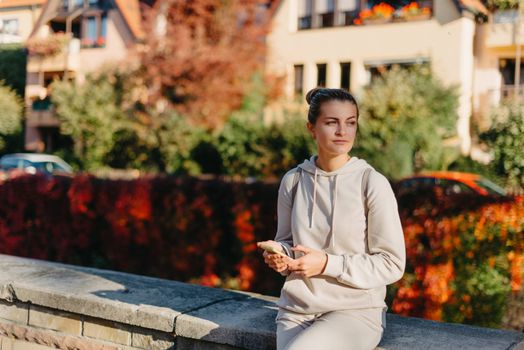 Young fashionable teenage girl with smartphone in park in autumn sitting at smiling. Trendy young woman in fall in park texting. Retouched, vibrant colors. Beautiful blonde teenage girl wearing casual modern autumn outfit sitting in park in autumn. Retouched, vibrant colors, brownish tones.