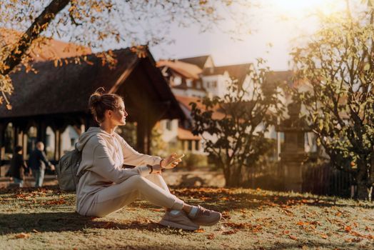 Young fashionable teenage girl with smartphone in park in autumn sitting at smiling. Trendy young woman in fall in park texting. Retouched, vibrant colors. Beautiful blonde teenage girl wearing casual modern autumn outfit sitting in park in autumn. Retouched, vibrant colors, brownish tones.