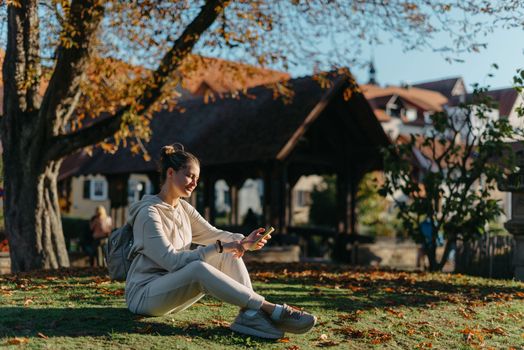 Young fashionable teenage girl with smartphone in park in autumn sitting at smiling. Trendy young woman in fall in park texting. Retouched, vibrant colors. Beautiful blonde teenage girl wearing casual modern autumn outfit sitting in park in autumn. Retouched, vibrant colors, brownish tones.