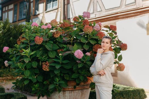 Attractive curly blonde woman walk on the city park street. Girl wear purple hoodie look happy and smiles. Woman make here me gesture standing near pink blooming bush flowers. Happy laughing girl