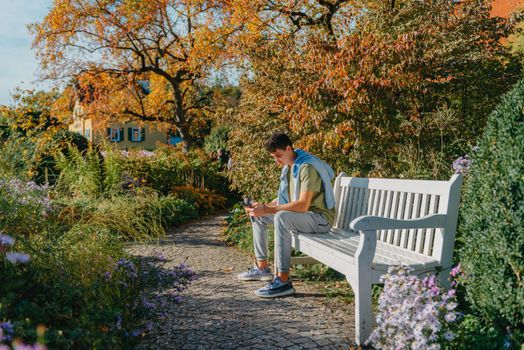 a teenager sits on a bench in the park drinks coffee from a thermo mug and looks into a phone. Portrait of handsome cheerful guy sitting on bench fresh air using device browsing media smm drinking latte urban outside outdoor.