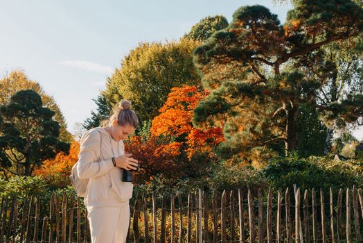 Beautiful young woman takes photos with a professional camera in autumn forest. Smiling girl enjoying autumn weather. Rest, relaxation, lifestyle concept. Young photographer takes pictures of autumn forest at sunset