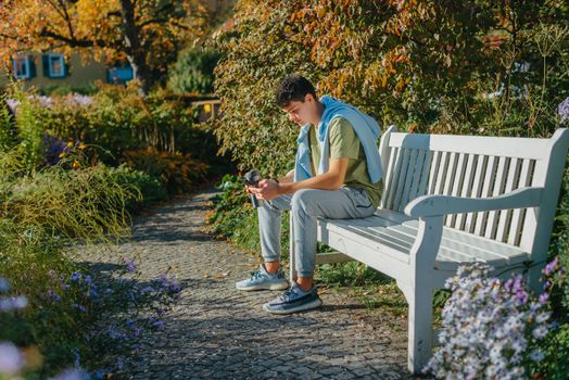 a teenager sits on a bench in the park drinks coffee from a thermo mug and looks into a phone. Portrait of handsome cheerful guy sitting on bench fresh air using device browsing media smm drinking latte urban outside outdoor.