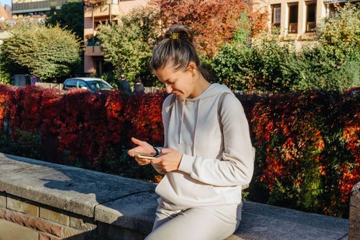 Young fashionable teenage girl with smartphone in park in autumn sitting at smiling. Trendy young woman in fall in park texting. Retouched, vibrant colors. Beautiful blonde teenage girl wearing casual modern autumn outfit sitting in park in autumn. Retouched, vibrant colors, brownish tones.