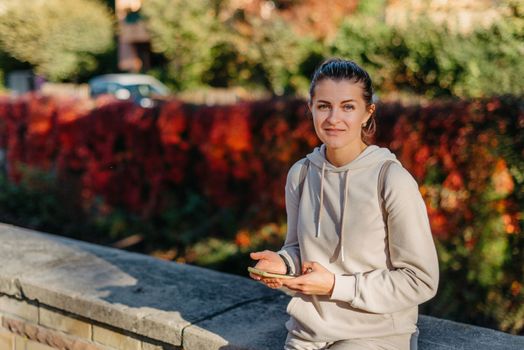 Young fashionable teenage girl with smartphone in park in autumn sitting at smiling. Trendy young woman in fall in park texting. Retouched, vibrant colors. Beautiful blonde teenage girl wearing casual modern autumn outfit sitting in park in autumn. Retouched, vibrant colors, brownish tones.