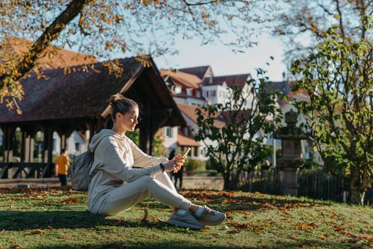 Young fashionable teenage girl with smartphone in park in autumn sitting at smiling. Trendy young woman in fall in park texting. Retouched, vibrant colors. Beautiful blonde teenage girl wearing casual modern autumn outfit sitting in park in autumn. Retouched, vibrant colors, brownish tones.