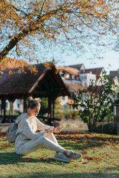 Young fashionable teenage girl with smartphone in park in autumn sitting at smiling. Trendy young woman in fall in park texting. Retouched, vibrant colors. Beautiful blonde teenage girl wearing casual modern autumn outfit sitting in park in autumn. Retouched, vibrant colors, brownish tones.