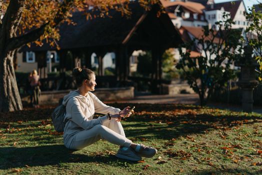 Young fashionable teenage girl with smartphone in park in autumn sitting at smiling. Trendy young woman in fall in park texting. Retouched, vibrant colors. Beautiful blonde teenage girl wearing casual modern autumn outfit sitting in park in autumn. Retouched, vibrant colors, brownish tones.